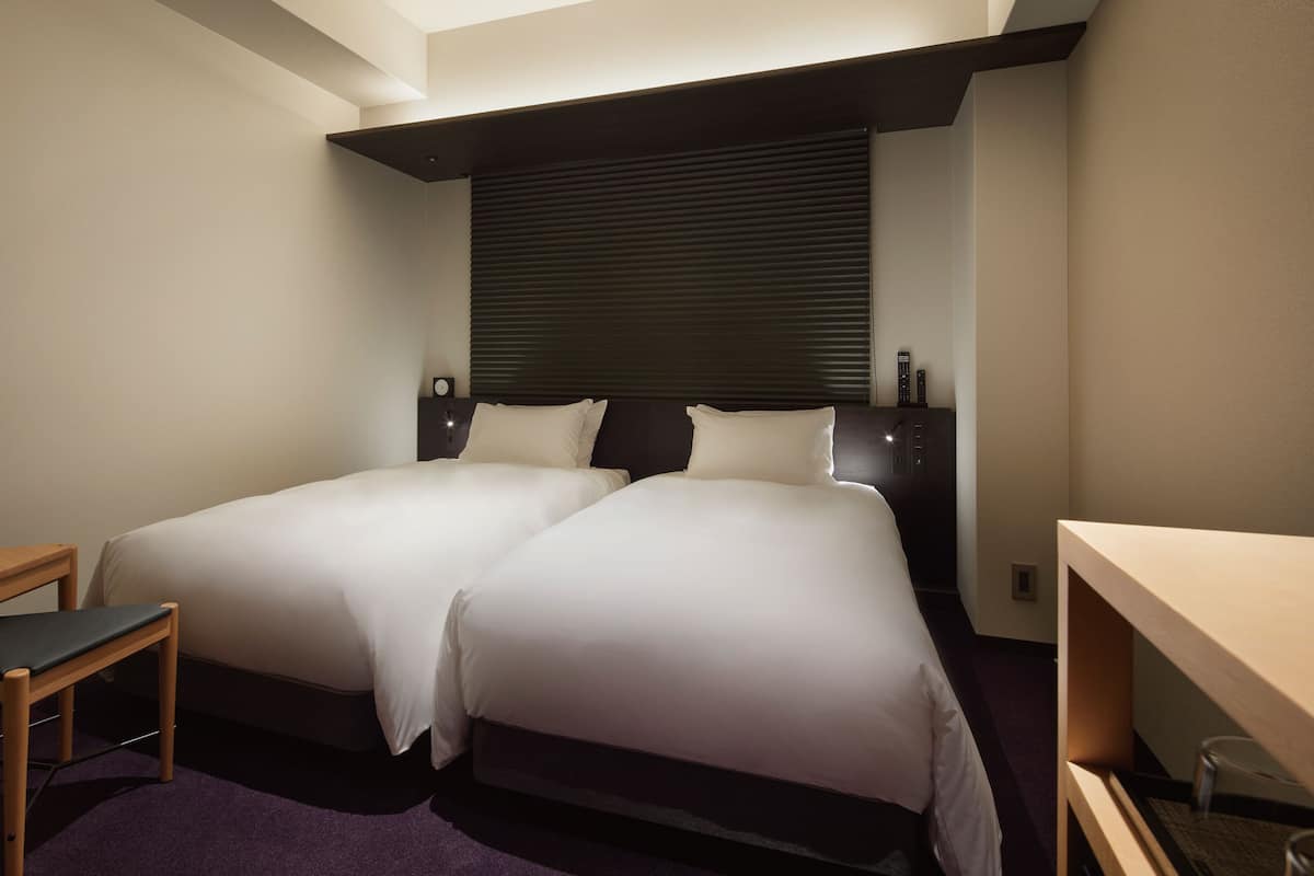 Une chambre d’hôtel avec deux lits, un bureau et une fenêtre avec des stores.