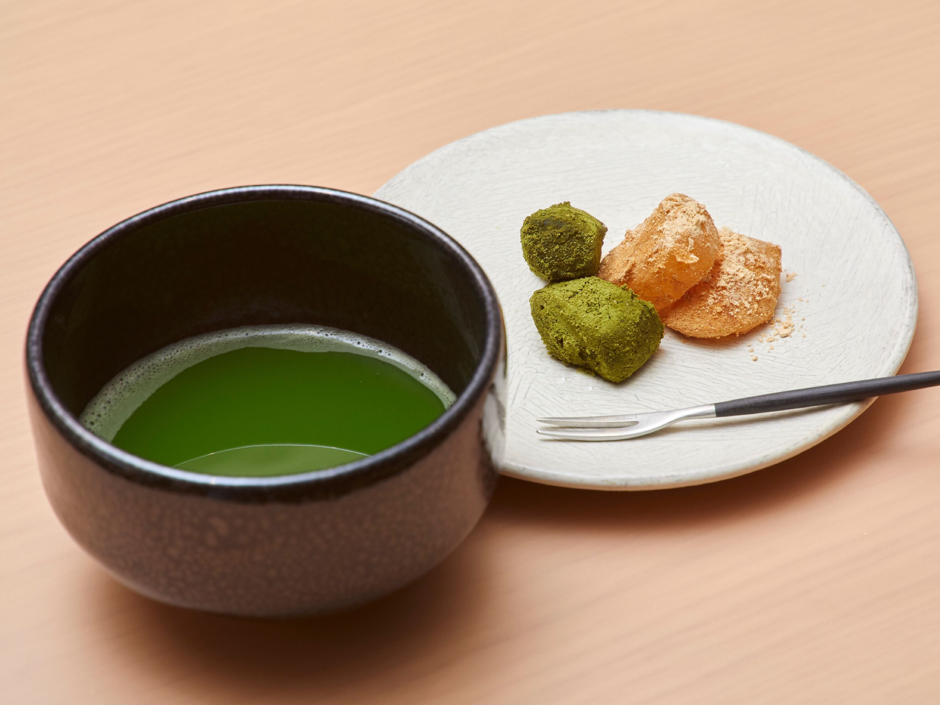 A dark ceramic cup filled with green liquid, a white ceramic plate with three green and one orange dessert, and a fork on the plate.