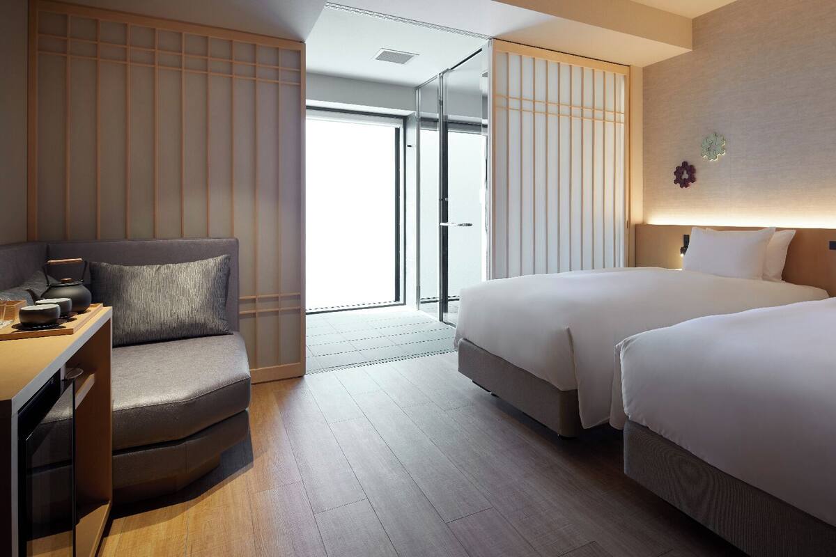 Une chambre d’hôtel avec un canapé, un bureau avec un service à thé, deux lits et une porte coulissante donnant sur un balcon.