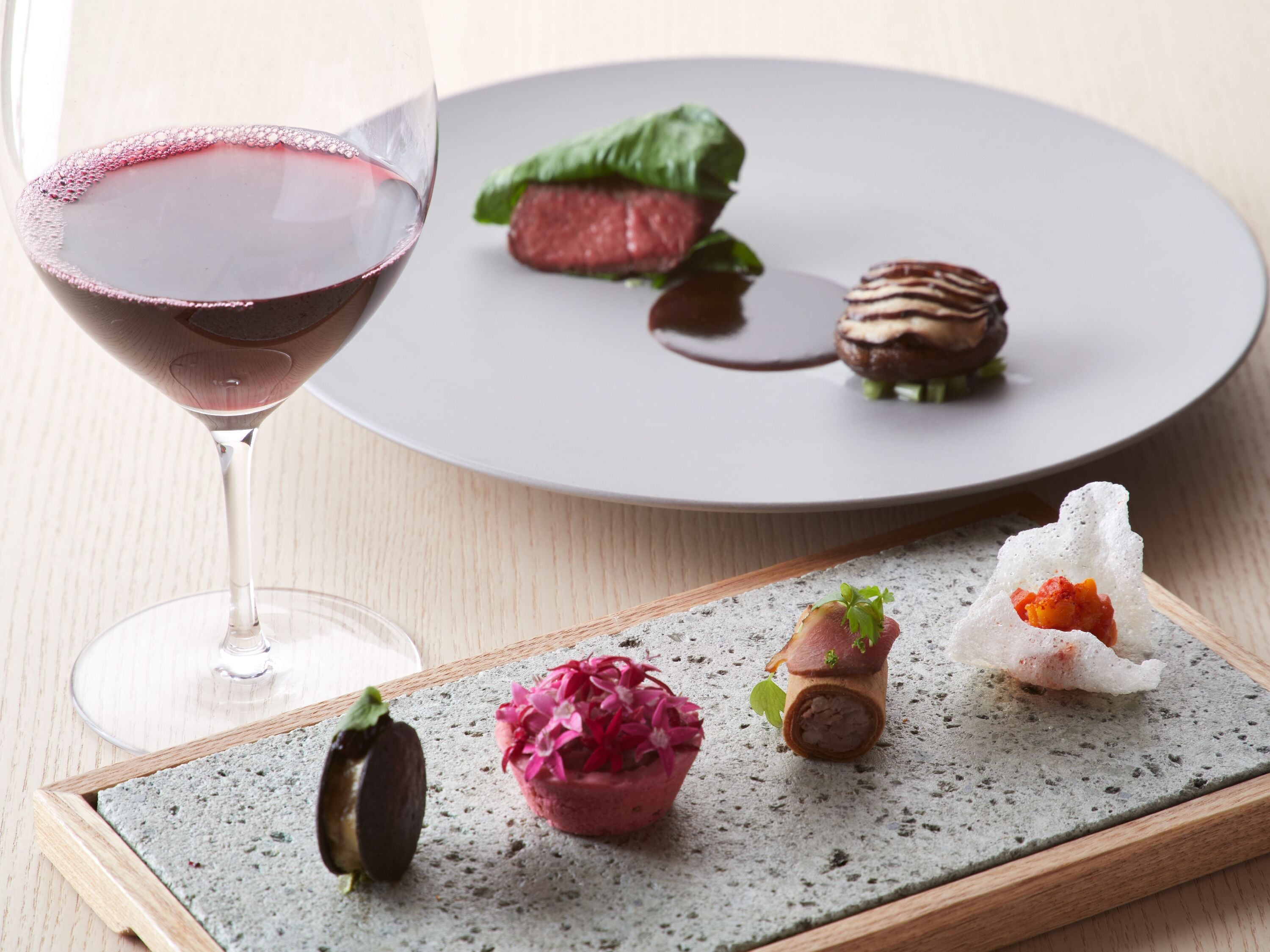 Uma mesa de jantar com uma taça de vinho, um prato com diversas opções de comida e um pequeno recipiente com uma flor.