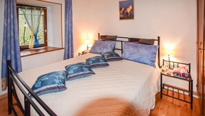 Ein ordentlich bezogenes Bett mit vielen Kissen und einem Kopfteil, ein Nachttisch mit Lampe und ein Fenster mit Vorhängen.