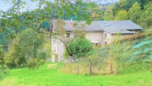 Ein Gebäude aus Stein mit Ziegeldach, umgeben von üppiger Vegetation und einem Zaun.