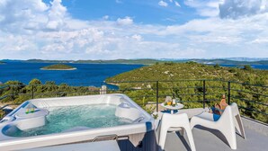 A hot tub with a table and chairs on a balcony overlooking a sea with islands and a mountainous landscape.