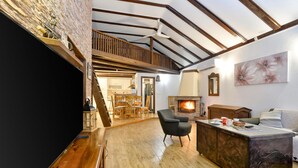 Amplia sala de estar con piso de madera, chimenea, comedor con mobiliario de madera y un altillo con vigas a la vista.