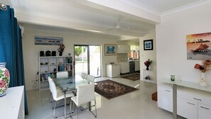 A modern kitchen with white cabinets, a dining area with a glass table and white chairs, and a living space with a large window and a ceiling fan. - 24 Lihs Street Elliott Heads (ELLIOTT HEADS)