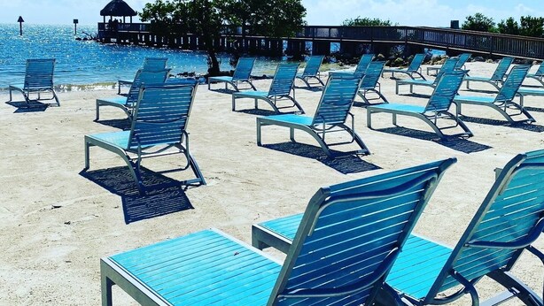 A beach with rows of blue lounge chairs and a wooden structure in the background. - 2BR - Key Largo Sunshine Getaway (Tavernier)