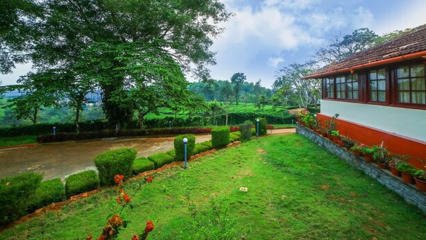 A lush green garden with a pathway leading to a building with red and white walls. - Deluxe Double Room In British Bungalow (Vythiri)