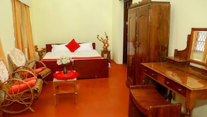 A bedroom with a bed, wooden wardrobe, wicker chairs, a small table with a vase, and a wooden desk. - Standard Double Room-wayanad (Vythiri)