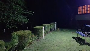 A nighttime outdoor area with a bench, trimmed hedges, and a building with illuminated windows. - Bungalow With Tea Garden View (Vythiri)