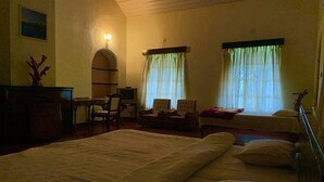 A room with a large bed, a desk with a chair, two windows with curtains, and a small table with a chair. - Bungalow With Tea Garden View (Vythiri)