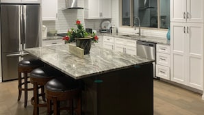 A modern kitchen with white cabinets, a stainless steel refrigerator, a marble island with stools, and a large window. - Golf Course Villa (East Wenatchee)