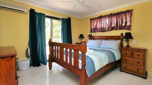 A bedroom with a wooden bed, two bedside tables with lamps, a chest of drawers, a window with curtains, and an air conditioning unit. - 36 Trevors Road Bargara (Bargara)