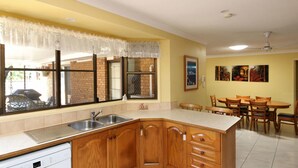 A kitchen with wooden cabinets, a stainless steel sink, and a dining area with wooden chairs and a table. - 36 Trevors Road Bargara (Bargara)