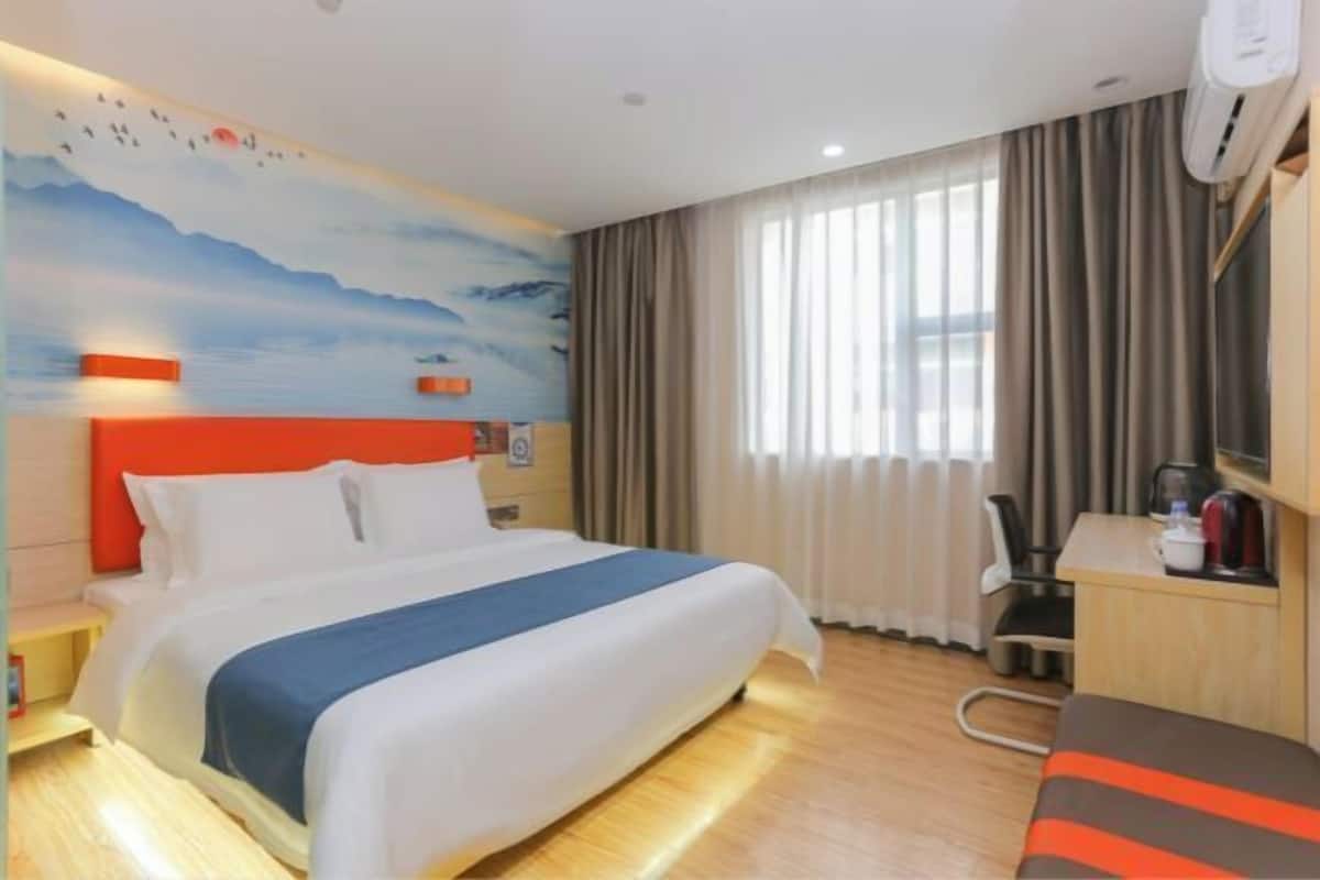 A hotel room with a bed, a desk, a chair, a radiator, and a window with curtains.