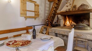 Ein rustikaler Essbereich mit Kamin aus Stein, Holzbalken und einem Tisch, gedeckt mit Tellern, Gläsern und einer Weinflasche.