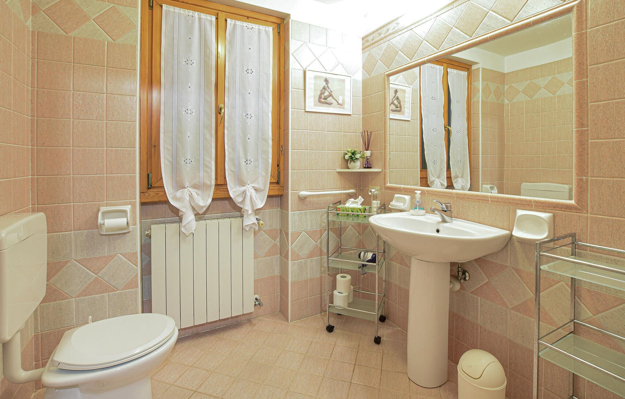 Een badkamer met toilet, wastafel en spiegel. Handdoeken hangen aan de muur, er is een radiator en een plank met toiletartikelen.