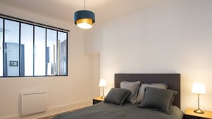 A modern bedroom with a large bed, two bedside lamps, a window with blinds, and a ceiling light. - Suite Napoléon III - 6.pers - downtown Charleville-Mézières (Charleville-Mézières)