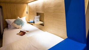Quarto pequeno e acolhedor com uma cama, um laptop numa prateleira e um livro na cama.