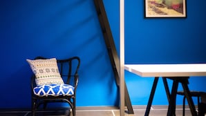 Quarto com parede azul, cadeira preta com almofadas estampadas, mesa branca e um pôster emoldurado na parede.