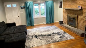 Un salon avec un canapé noir, des rideaux turquoise, une cheminée et une vue sur la verdure à l’extérieur.