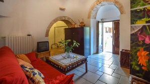 Ein Wohnzimmer mit einem roten Sofa, einem kleinen Tisch mit weißer Spitzenleinentuch, einem Fernseher und einer Pflanze. Zwei Bogenfenster führen in einen anderen Raum.