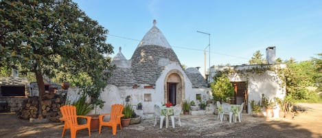 Un edificio rústico de piedra con techo cónico, zona de estar al aire libre con sillas de plástico y macetas con plantas.