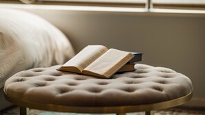 A round, tufted ottoman with an open book on it, placed on a glass table in a room with a large window and a bed in the background. - Swwitch House Central Tokyo 7 (Tokyo)