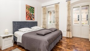 A bedroom with a large bed, a nightstand, a lamp, a painting, and a view through a window with curtains. - Cozy apartment in Volosko with WiFi (Volosko)
