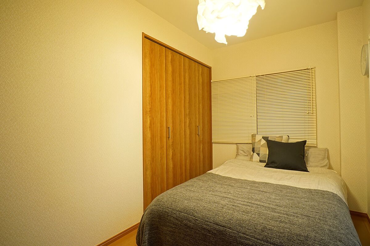 A bedroom with a bed, a wooden wardrobe, a window with blinds, and a light fixture.