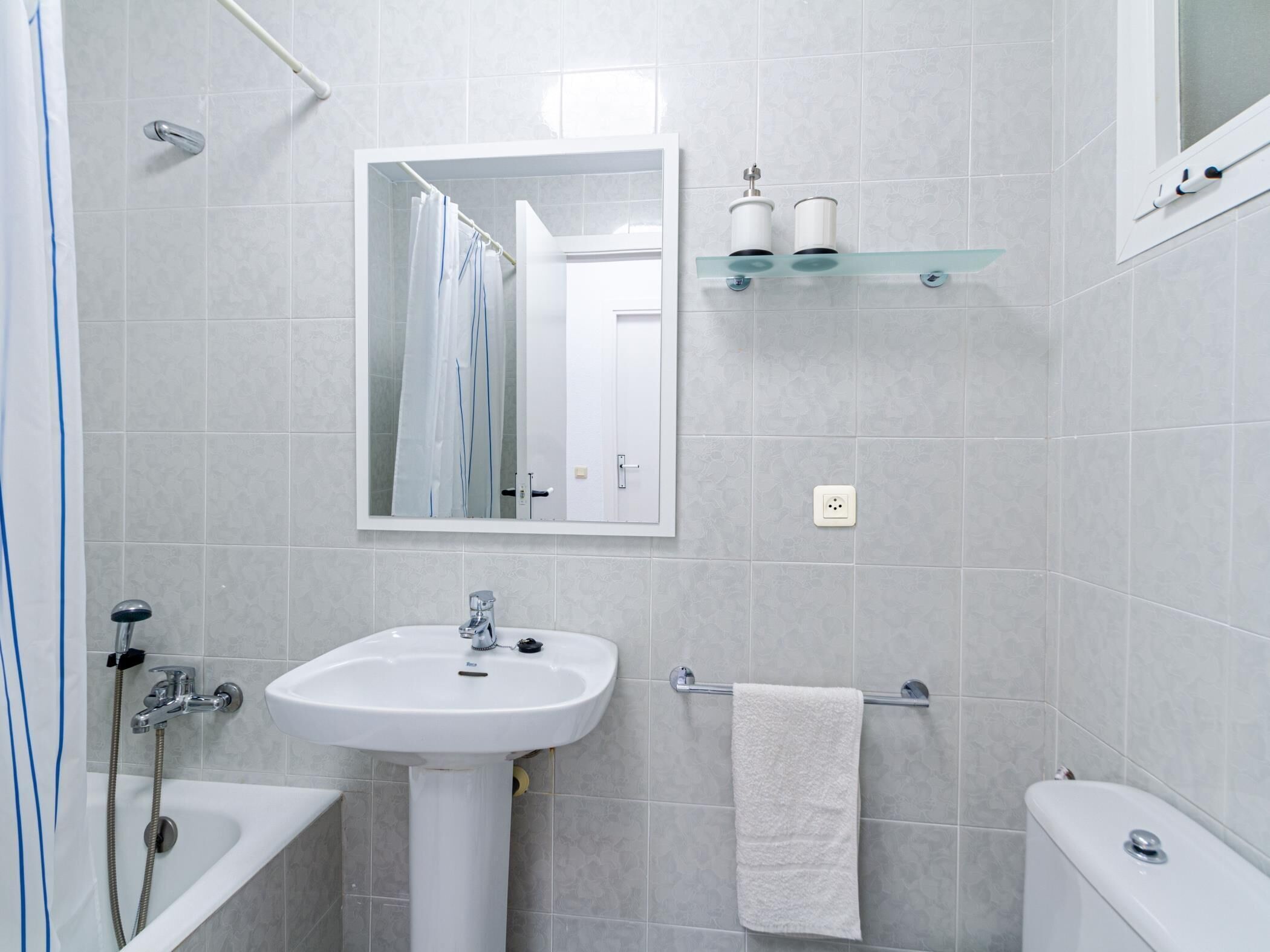 Een badkamer met een witte wastafel, een spiegel, een handdoekhouder, een glazen plank met toiletartikelen en een douche.