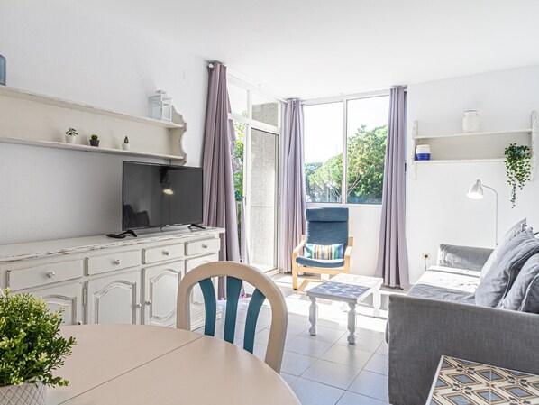 A modern kitchen with a dining table, a chair, and a sofa. There is a TV on a cabinet, and a window with curtains.