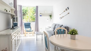 Un salon lumineux et moderne, avec un canapé blanc, un fauteuil bleu, une table à manger et une grande fenêtre donnant sur un paysage verdoyant.