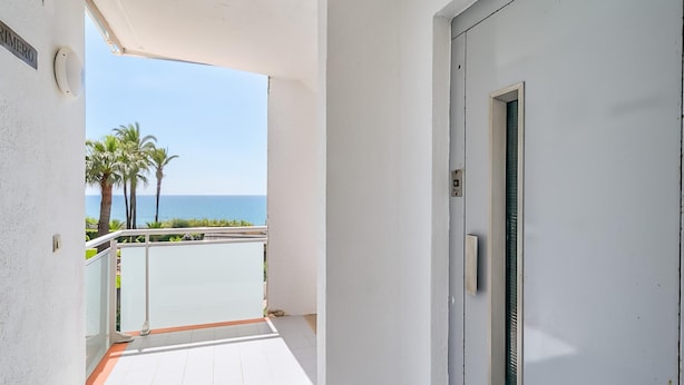 Un couloir avec une porte blanche, un petit balcon donnant sur la mer et les palmiers, et un sol carrelé.