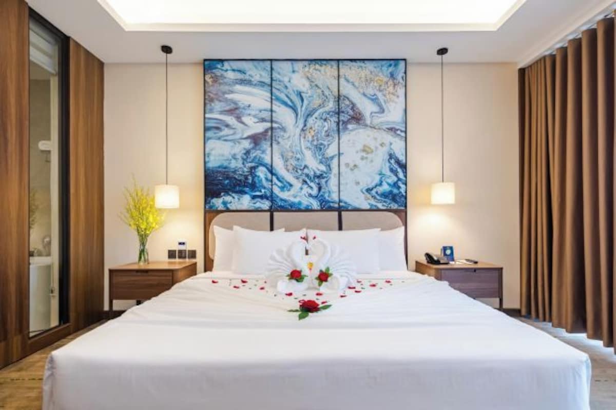 A hotel room with a large bed, a painting above the bed, and a nightstand with a lamp.