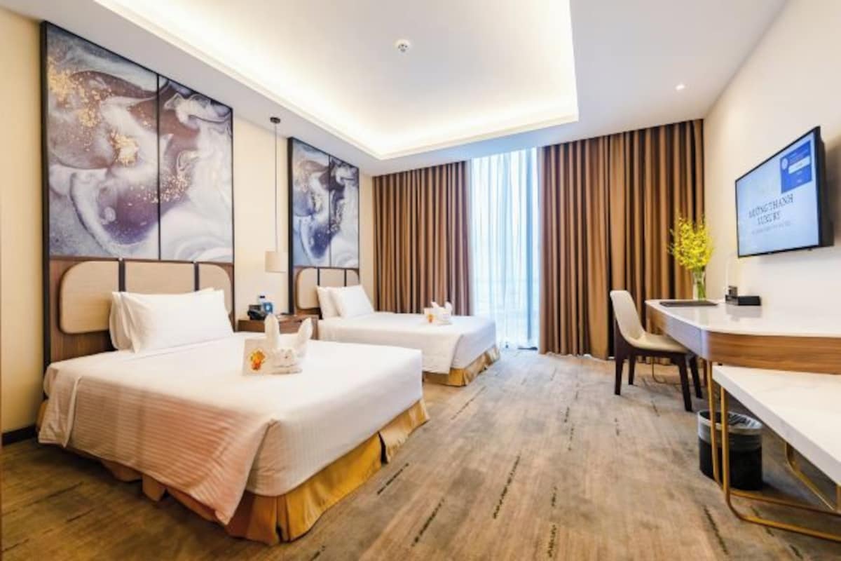 A hotel room with two beds, a desk, and a chair. There are abstract paintings on the walls and a television on the wall.