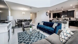 Ein modernes Wohnzimmer mit einem blauen Sofa, grauen Sesseln und einem Couchtisch. Es gibt einen Essbereich mit einer Chandelier und eine Küche mit dunklen Schränken und einer Mikrowelle.