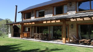 A two-story log cabin with a stone fireplace, outdoor seating, and a large window overlooking a grassy area. - Alhue - Casa en el Golf (San Carlos de Bariloche)