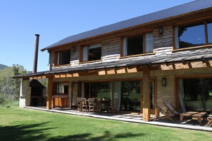 Une cabane en rondins à deux étages, dotée d’une cheminée en pierre, d’un espace salon extérieur et d’une grande fenêtre donnant sur une zone verdoyante.