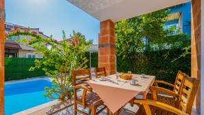 Une terrasse avec une table et des chaises, une piscine et une vue sur les maisons et la verdure.