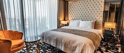 Una habitación de hotel con una cama grande, un cabecero tapizado, una mesa redonda, un sillón de cuero y una alfombra estampada.