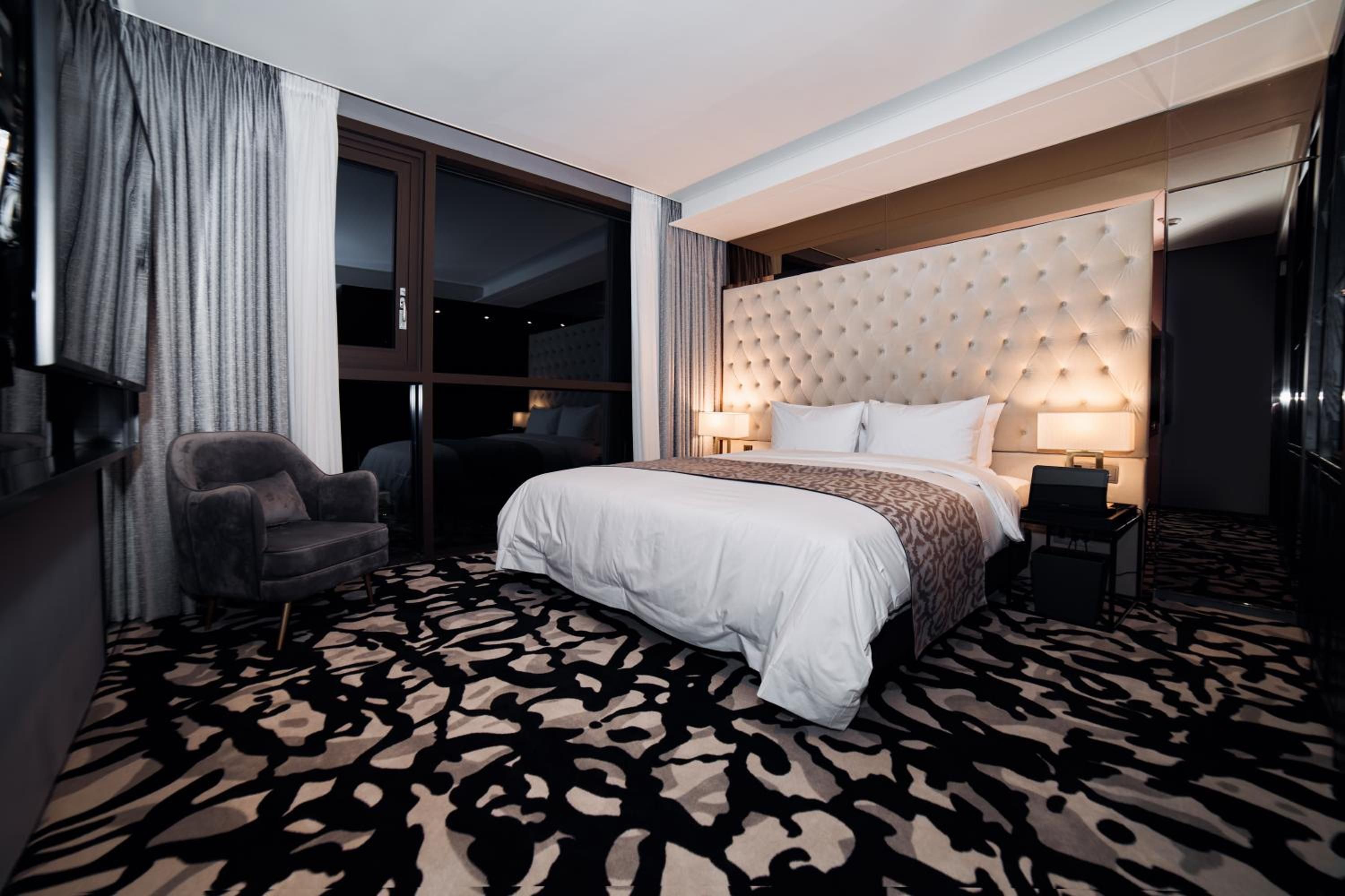 A hotel room with a large bed, a chair, a desk with a laptop, and a large window.