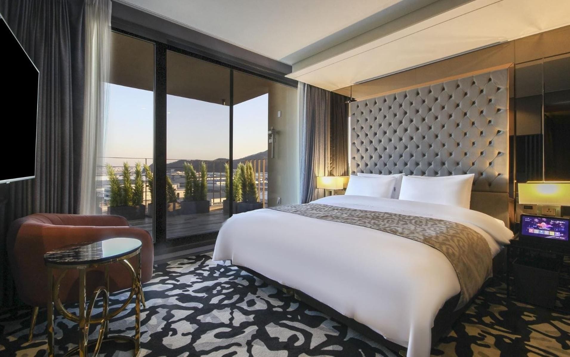 A hotel room with a large bed, a seating area with a table, a TV, and a view of the cityscape.