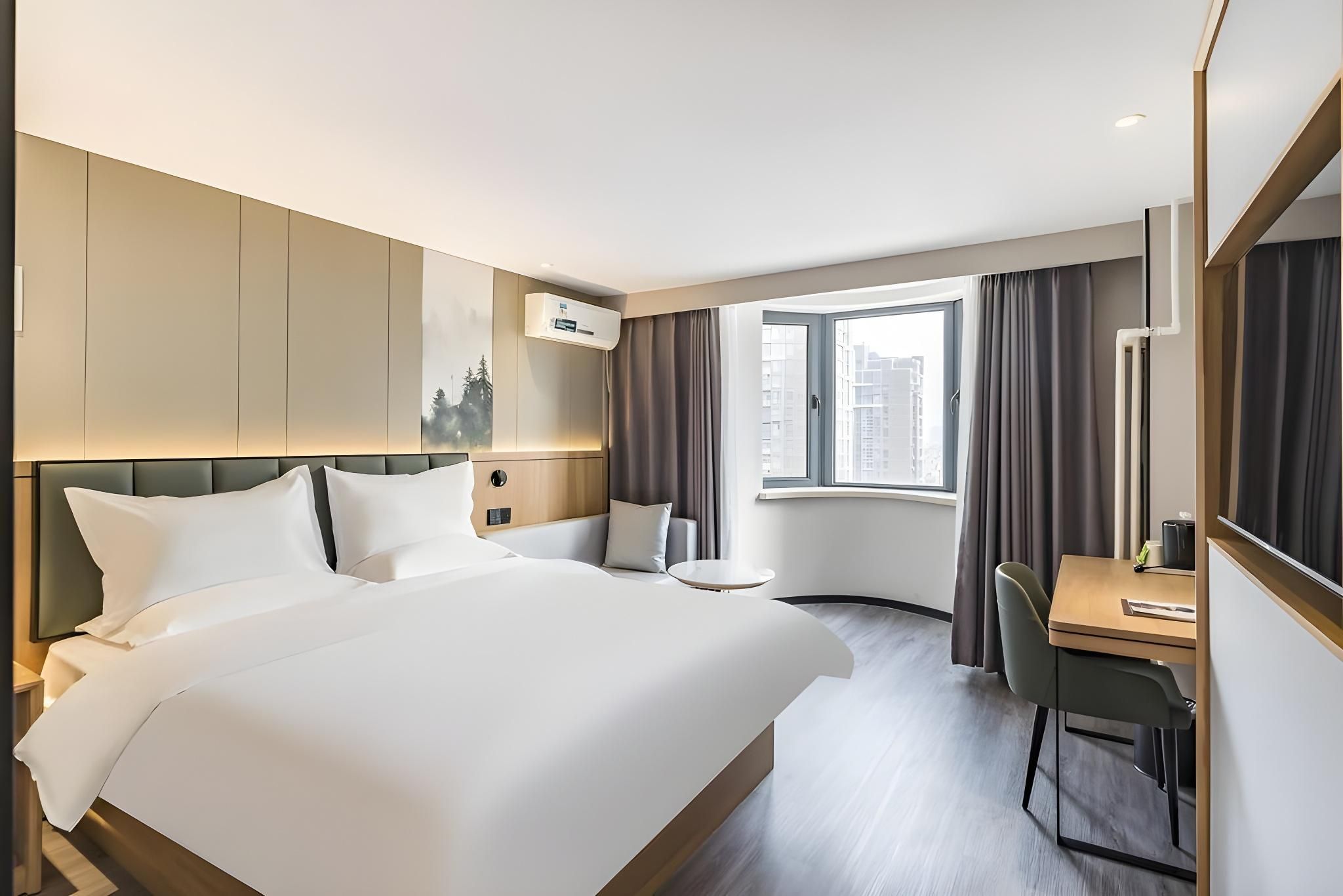 A hotel room with a large bed, a desk, a chair, a window with curtains, and a wall-mounted air conditioning unit.