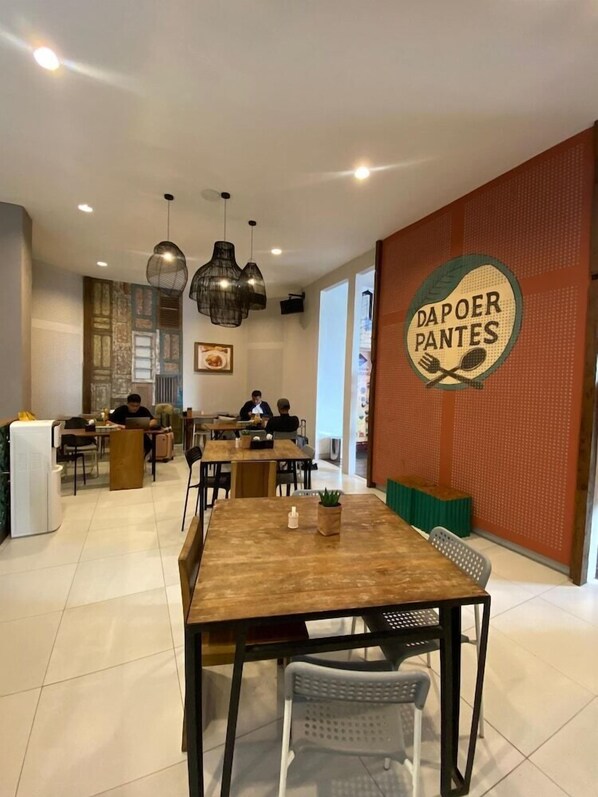 A modern dining area with wooden tables and chairs, a wall-mounted sign reading "Dapoer Pantes," and a seating area with a green bench. - Hotel Pantes Simpang Lima Semarang (Semarang)