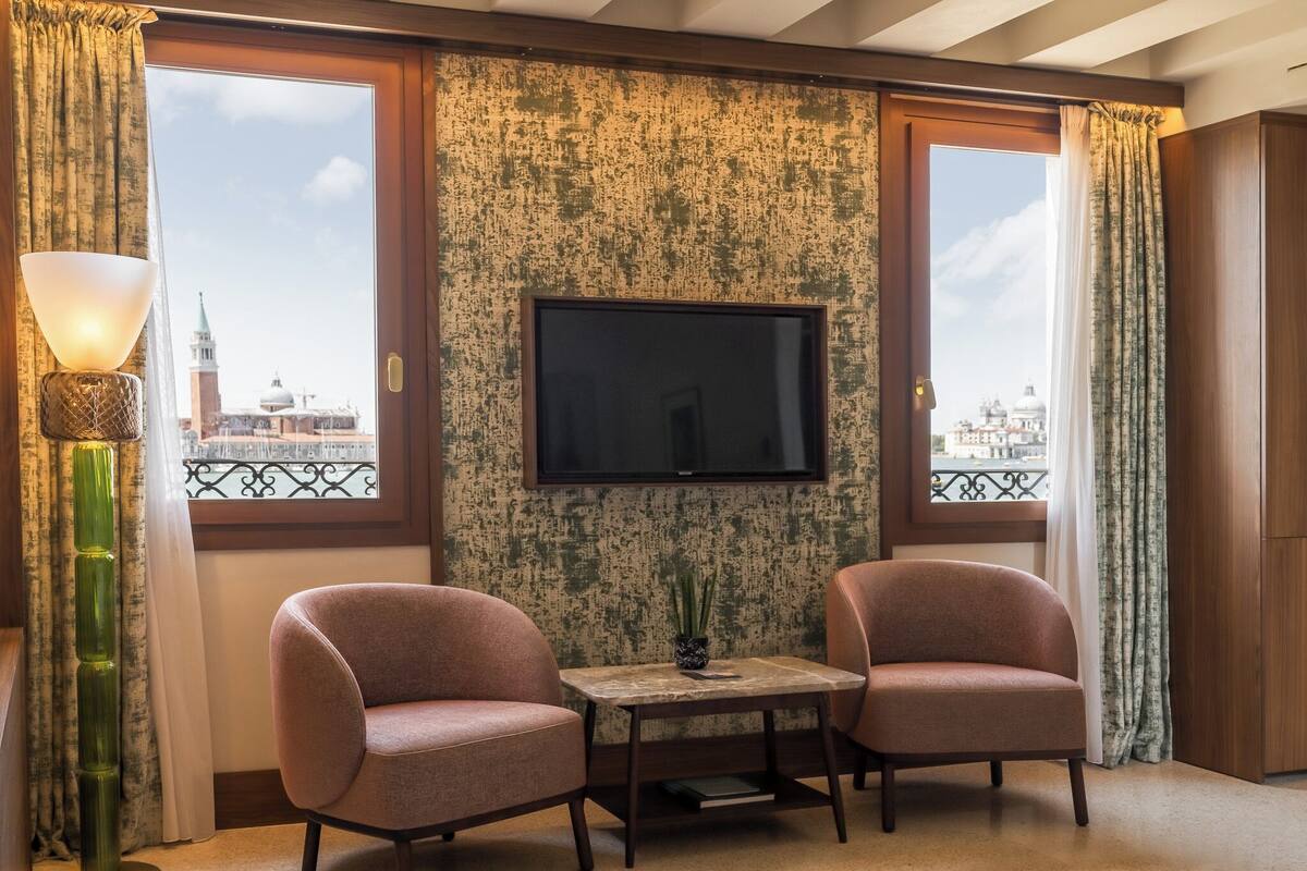 A room with two chairs, a small table, a TV on the wall, and a view of a cityscape through large windows.
