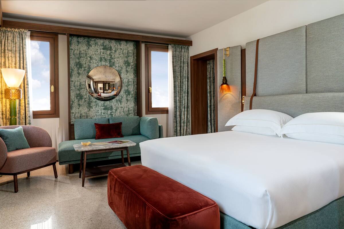 A hotel room with a bed, a sofa, a small table, a red ottoman, a mirror, and curtains.