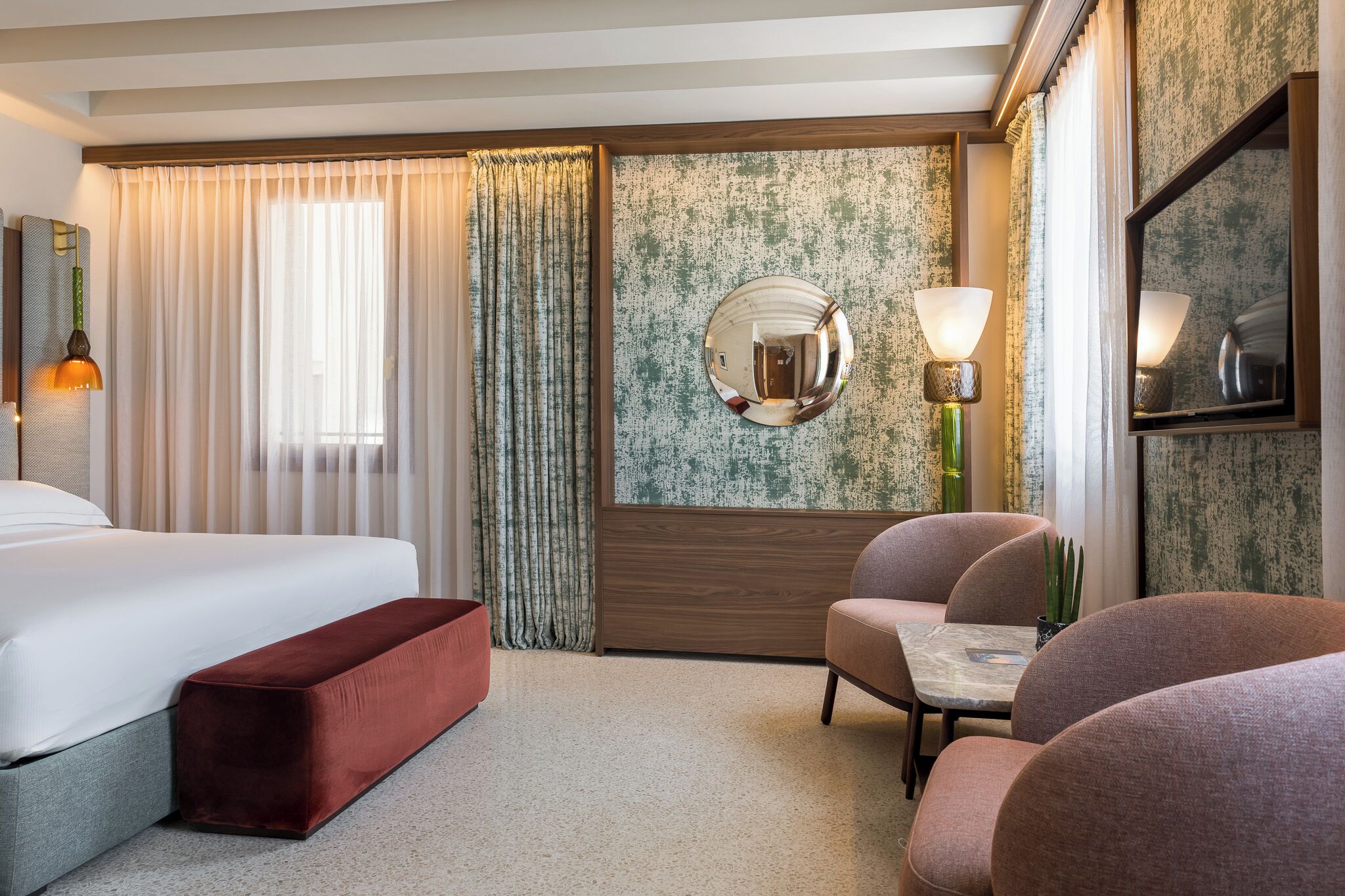 A hotel room with a bed, a red bench, two chairs, a round mirror, and two wall-mounted lamps.