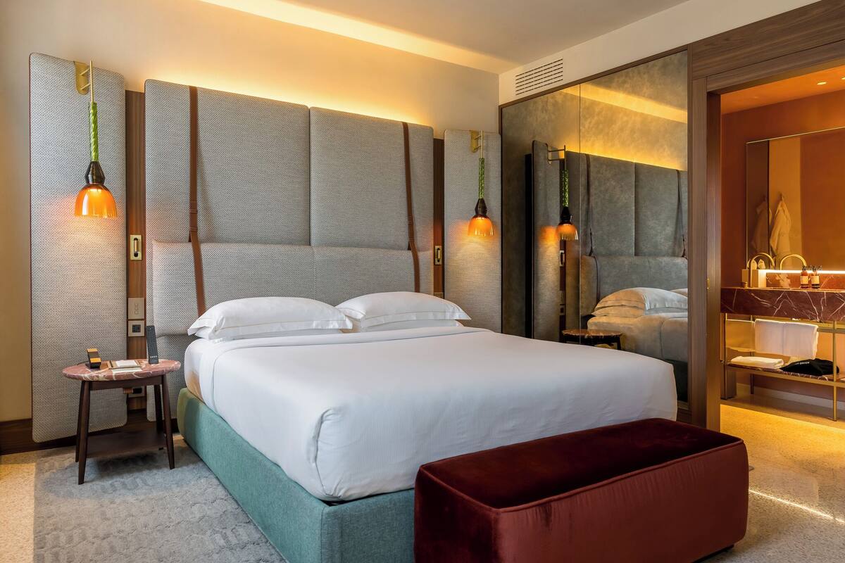 A modern hotel room with a large bed, a bedside table, a red ottoman, and a bathroom with a mirror and sink.