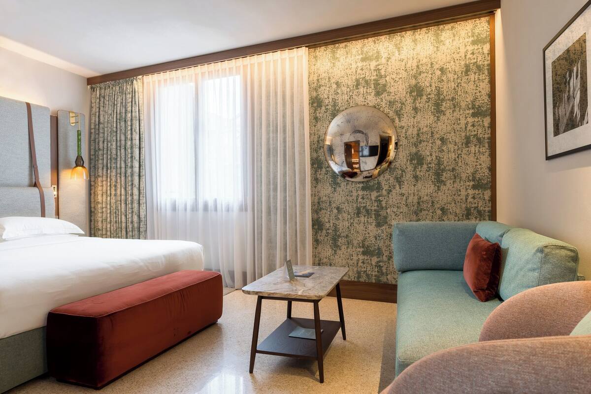 A hotel room with a bed, a sofa, a small table, and a wall-mounted mirror.