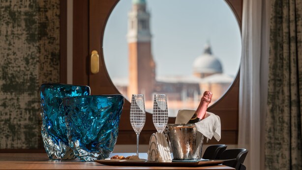 A table with a blue glass bowl, two champagne flutes, a silver bucket, and a champagne bottle. A view of a building with a dome and a tower is visible through a round window. - Ca' di Dio - VRetreats, SLH Collection (Venice)